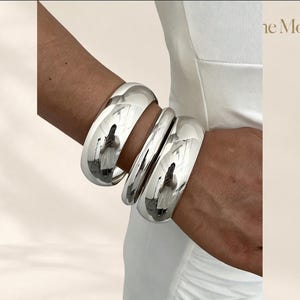 Könnte beinhalten: Drei polierte Armreifen aus silberfarbenem Metall mit einer glatten, abgerundeten Oberfläche. Die Armbänder haben ein hochglänzendes, reflektierendes Finish. Der Text "the Moft" ist in der oberen rechten Ecke zu sehen, zusammen mit einem kleinen sternförmigen Symbol.
