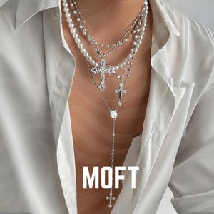 Pode incluir: Colar de prata em camadas com pérolas e pingentes de cruz, usado sobre uma camisa branca. A palavra "MOFT" é exibida em letras brancas na parte inferior da imagem.