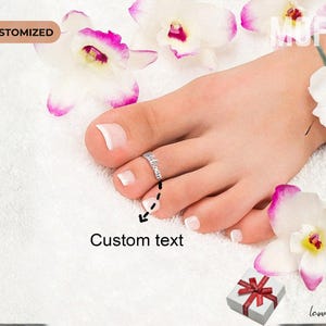 May include: A silver toe ring with custom text is worn on a foot with a white pedicure. White and purple orchids and a small gift box with a red ribbon are arranged around the foot. The word "CUSTOMIZED" is in the upper left corner.