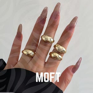 Puede incluir: Una mano que muestra cinco anillos dorados con un diseño suave y redondeado. Los anillos se usan en varios dedos. La palabra "MOFT" se muestra en letras blancas. El fondo es de color claro.
