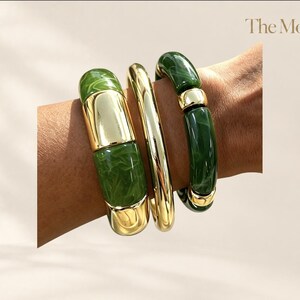 Könnte beinhalten: Ein Set aus drei Armbändern mit abwechselnden Segmenten aus poliertem goldfarbenem Metall und marmoriertem waldgrünem Harz. Das Design umfasst einen massiven goldfarbenen Armreif und zwei Armbänder mit zylindrischem grünem Harz und Goldakzenten.