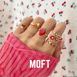 May include: A hand wearing several gold-toned rings with fruit and candy designs. The rings include a cherry, a strawberry, and a candy ring. The hand is wearing a pink sweater. The background is a white fabric with a cherry pattern.