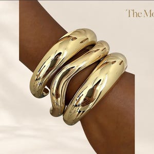 May include: Three polished gold-tone metal bangles with a smooth, rounded, and thick tubular design. The bracelets feature a high-shine reflective surface and are stacked together. The text "The Moft" is visible in the top right corner.
