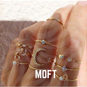 Può includere: Una collezione di anelli dorati con vari design, tra cui una luna crescente, stelle, cuori e accenti floreali. Gli anelli sono esposti su dita, mostrando i loro delicati dettagli e trame. La parola "MOFT" è visualizzata in testo bianco.