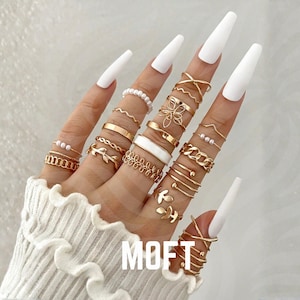 Peut inclure: Ensemble de bagues dorées de différents modèles, dont des anneaux, des chaînes et des motifs floraux. Certaines bagues présentent des perles ou de l'émail blanc. Les bagues sont portées sur des doigts aux longs ongles blancs. Le mot "MOFT" est visible.