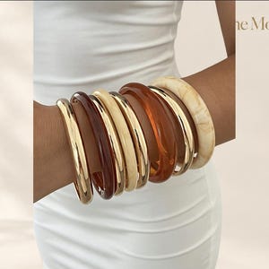 Op de afbeelding: Een stapel van acht armbanden met een mix van gepolijst goudkleurig metaal, doorschijnende amberkleurige hars, effen crèmekleurige hars en een gemarmerde beige harsafwerking. De armbanden worden samen gedragen en tonen gladde texturen.