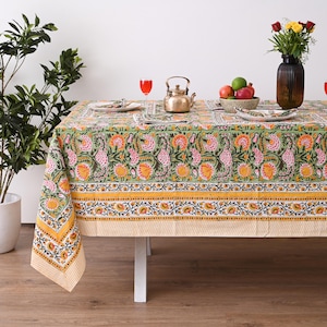May include: A rectangular green tablecloth with a floral pattern in pink and orange. The tablecloth has a yellow border with a floral design and is set on a white table. A vase of flowers and a teapot are on the table.