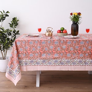 Può includere: Una tovaglia color corallo con un motivo floreale con accenti blu e bianchi. La tavola è apparecchiata con piatti, bicchieri e un vaso di fiori. Una teiera in ottone e una ciotola di frutta completano la scena.
