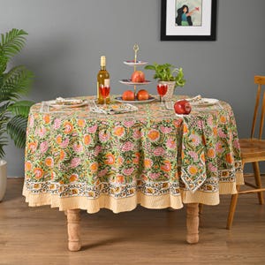 May include: A round table set for dining, featuring a green floral tablecloth with orange and pink accents. The table is set with plates, napkins, and a tiered serving dish. A bottle of wine and glasses are also present.
