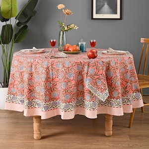 May include: A round coral tablecloth with a floral design in blue and green. The table is set with wine glasses, plates, and fruit. The tablecloth is on a wooden table, creating a dining setting.