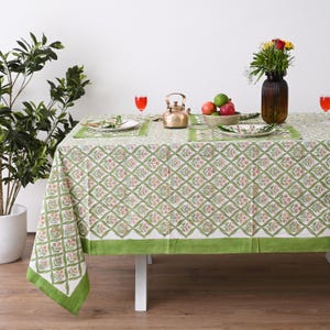 May include: A green and white floral patterned tablecloth with matching placemats and napkins. The table is set with a brass teapot, a bowl of fruit, and a vase of flowers. The tablecloth has a green border.