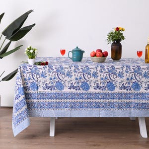 Könnte beinhalten: Eine weiße Tischdecke mit blauem Blumenmuster und gestreiftem Rand. Der Tisch ist mit einer Teekanne, einer Schüssel mit Äpfeln, einer Vase mit Blumen und zwei Gläsern roter Flüssigkeit gedeckt. Eine Pflanze befindet sich im Hintergrund.