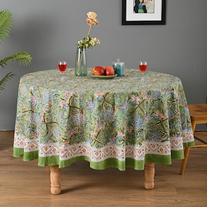 Może przedstawiać: Okrągły zielony obrus z kwiatowym wzorem, ustawiony na drewnianym stole. Obrus ma szczegółowy wzór z niebieskimi, pomarańczowymi i białymi akcentami. Na stole wazon z kwiatami, dwa kieliszki czerwonego wina i talerz z jabłkami.