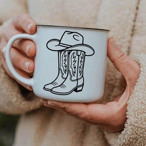 May include: White ceramic mug with a black rim featuring a black illustration of a cowboy hat and cowboy boots. The boots have decorative stitching. The mug is held by a person.
