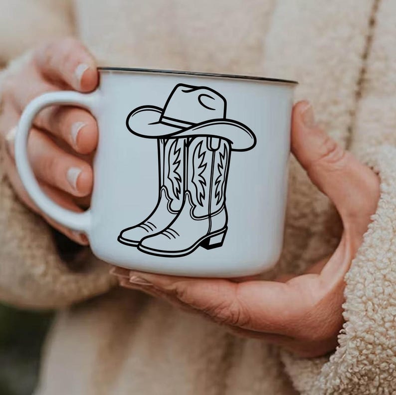 May include: White ceramic mug with a black rim featuring a black illustration of a cowboy hat and cowboy boots. The boots have decorative stitching. The mug is held by a person.
