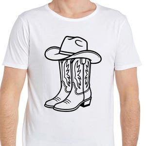 May include: White t-shirt featuring a black and white graphic of a cowboy hat resting on top of a pair of cowboy boots. The boots have decorative stitching. The design is centered on the shirt.