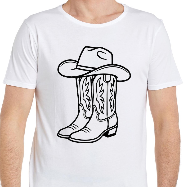 May include: White t-shirt featuring a black and white graphic of a cowboy hat resting on top of a pair of cowboy boots. The boots have decorative stitching. The design is centered on the shirt.