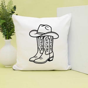May include: White square pillow featuring a black and white illustration of a cowboy hat resting on top of a pair of cowboy boots. The boots have decorative stitching.