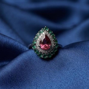 Può includere: Un anello in argento con una pietra preziosa rosa centrale a forma di goccia, circondata da un alone di piccole pietre bianche e da un bordo esterno di pietre verdi rotonde. L'anello è montato su una fascia metallica dal design complesso.