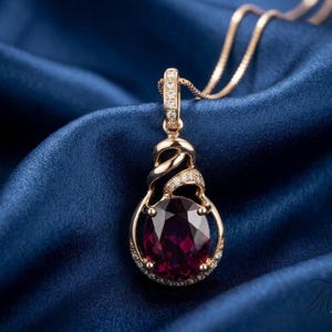 May include: A gold-colored pendant necklace with a large, oval, deep red gemstone. The pendant is surrounded by small, clear gemstones and has a decorative, twisted design at the top. The necklace is displayed on a blue satin fabric.