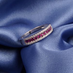 May include: A silver ring with a row of vibrant red gemstones set between two rows of smaller, clear gemstones. The ring is displayed on a blue satin fabric, highlighting its sparkle and design.