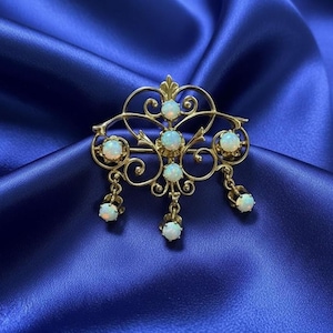 May include: A vintage gold-toned brooch with opal gemstones. The brooch features an ornate design with a central cluster of opals and smaller opals suspended from delicate chains. The brooch is set against a blue satin background.