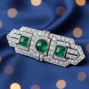 Op de afbeelding: Zilverkleurige broche met een rechthoekig ontwerp, met drie groene smaragden en talrijke kleine, sprankelende diamanten. De broche heeft een Art Deco-stijl en staat tegen een blauwe achtergrond.
