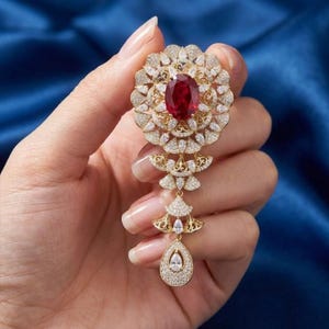 May include: A gold-toned brooch featuring a large oval ruby center stone surrounded by a halo of smaller clear gemstones. The brooch has a floral design with a dangling teardrop-shaped pendant.