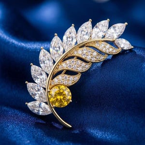 May include: A gold-toned brooch in a leaf design, featuring clear and yellow gemstones. The brooch has a curved stem with clear, marquise-cut stones on one side and smaller, round stones on the other. A large, round yellow stone is at the base.