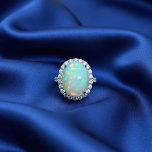 Peut inclure: Bague ovale en opale, entourée d'un halo de petites pierres précieuses transparentes. L'opale a une base blanc laiteux avec des reflets verts et bleus. La bague est sertie sur un anneau argenté, posée sur un tissu de satin bleu.