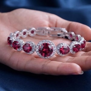 May include: A silver-toned bracelet featuring a series of round, deep red gemstones. Each stone is surrounded by a halo of small, clear, sparkling crystals. The bracelet includes a secure metal clasp mechanism and a flexible link design.