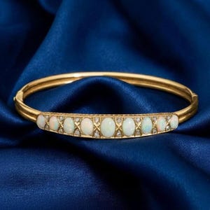 May include: A gold bangle bracelet featuring oval opals and small diamonds. The bracelet is displayed on a dark blue satin backdrop, accentuating the warm hues and detailed craftsmanship of the jewellery.