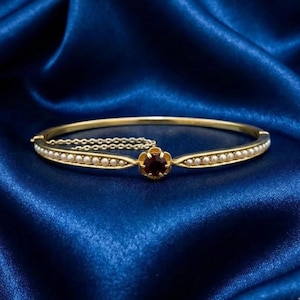 Puede incluir: Una pulsera dorada con una piedra preciosa roja oscura central en un engaste floral. La pulsera está adornada con pequeñas perlas blancas y descansa sobre una superficie de satén azul. Una fina cadena asegura el cierre.