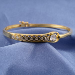 Könnte beinhalten: Ein goldfarbenes Armreif-Armband mit einer Reihe kleiner, klarer Edelsteine und einem größeren, runden Edelstein. Das Armband ist vor einem blauen Hintergrund platziert, der seine polierte Oberfläche und sein elegantes Design hervorhebt.