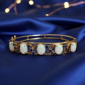 Puede incluir: Una pulsera de brazalete dorada con piedras preciosas blancas ovaladas y pequeñas piedras preciosas azul oscuro. La pulsera está colocada sobre un fondo azul oscuro, resaltando el diseño de la joya.