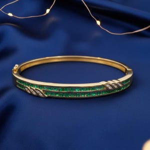 May include: A gold bangle bracelet featuring a row of emerald-cut green gemstones and small diamonds. The bracelet is set against a dark blue background, with decorative lights.