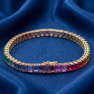 Puede incluir: Una pulsera dorada adornada con una serie de piedras preciosas rectangulares multicolores. Las piedras preciosas muestran un espectro de colores, incluyendo rojo, naranja, amarillo, verde, azul y morado. La pulsera está sobre un fondo azul oscuro.