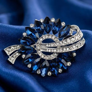 Puede incluir: Broche plateado con diseño floral. Presenta gemas ovales de color azul oscuro y cristales transparentes. El broche está sobre un fondo de satén azul, creando una apariencia lujosa.