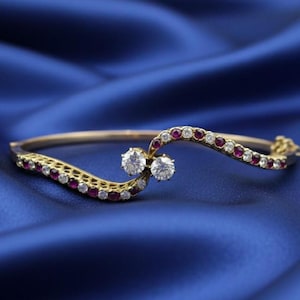 May include: A gold bracelet with a wave design, set with clear and red gemstones. Two large, clear gemstones are centered on the bracelet. The bracelet is displayed on a blue satin background.