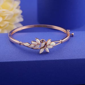 Puede incluir: Pulsera de oro rosa con diseño floral. La pulsera presenta piedras blancas similares al ópalo dispuestas en un delicado patrón curvo. La pulsera se exhibe sobre una superficie azul, con una flor amarilla en el fondo.