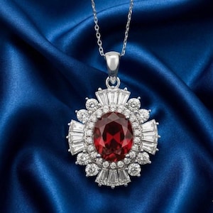 Può includere: Una collana d'argento con un pendente con una grande gemma ovale rosso intenso, circondata da gemme più piccole, chiare, taglio baguette e rotonde. Il pendente è appeso a una delicata catena d'argento su uno sfondo blu.