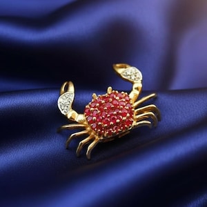 May include: A gold crab brooch featuring a body covered in small red gemstones. The crab's claws have clear, sparkling details. The brooch is set against a deep blue, silky background.