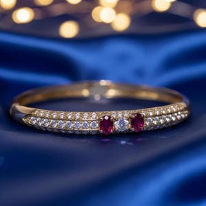 May include: A gold bangle bracelet featuring a row of small, clear gemstones and two oval-cut, red gemstones. The bracelet is set against a blue background with blurred, golden lights.