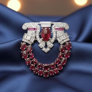 Può includere: Spilla argentata con un rubino ovale centrale e un design circostante di rubini più piccoli e cristalli trasparenti. La spilla ha un design simmetrico con una base curva e una parte superiore rettangolare, su sfondo blu.