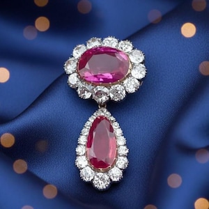 Puede incluir: Un lujoso broche con un gran rubí ovalado rodeado por un halo de diamantes. Un colgante de rubí en forma de pera cuelga debajo, también rodeado de diamantes. La joya está engastada sobre un fondo de satén azul oscuro.