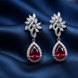 May include: A pair of silver-toned drop earrings featuring a pear-shaped red gemstone at the base, surrounded by a halo of small clear stones. Above the teardrop, a cluster of marquise-cut clear stones forms an intricate, leafy design.
