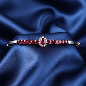 May include: A silver bracelet with a central oval ruby surrounded by small pearls. The bracelet features a row of small, round rubies set along the band. The background is a deep blue fabric.
