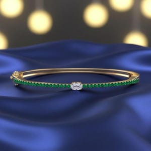 Könnte beinhalten: Ein goldener Armreif mit einer Reihe kleiner, runder grüner Edelsteine und einem einzelnen, rechteckigen, klaren Edelstein in der Mitte. Das Armband ist vor einem dunkelblauen, seidigen Hintergrund platziert.