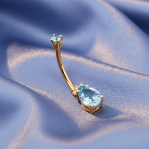 Puede incluir: Un piercing para el ombligo de color dorado con un diseño de barra curva. El anillo presenta dos piedras preciosas de color azul claro, una en forma de lágrima y otra redonda, con pequeñas piedras de acento transparentes. La joya está sobre un fondo azul.