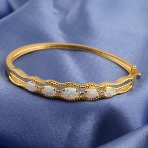 Puede incluir: Un brazalete dorado con cinco ópalos ovalados y pequeñas piedras de acento. El brazalete tiene un diseño decorativo ondulado a lo largo de los bordes. El cierre es visible en el lado derecho, sobre un fondo de tela azul.